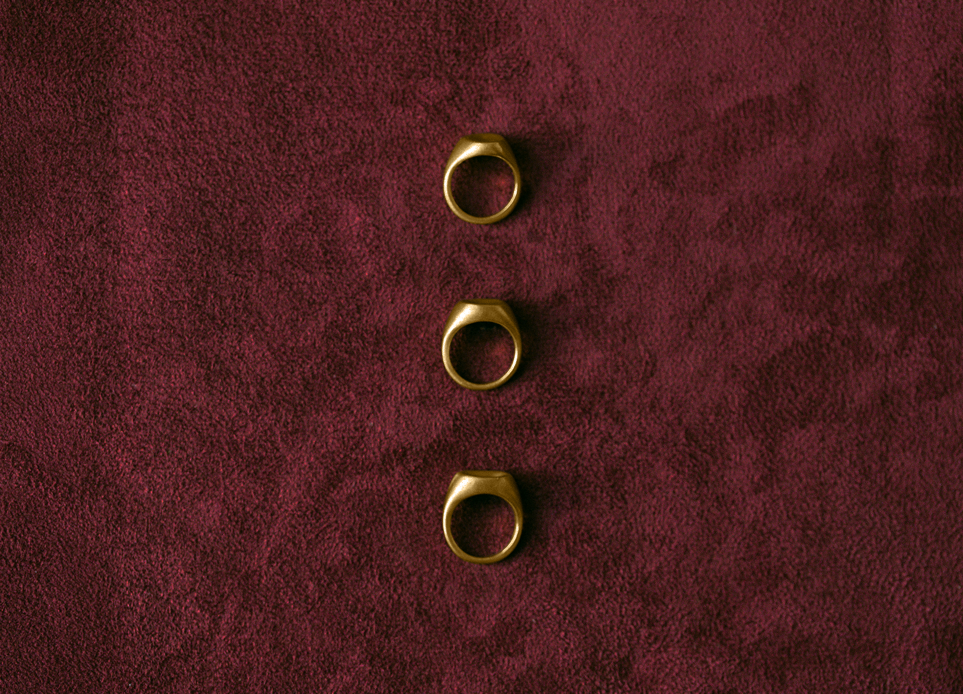 Three sculptural signet rings in 14k gold by dear_ jewelry arranged vertically on textured burgundy fabric.